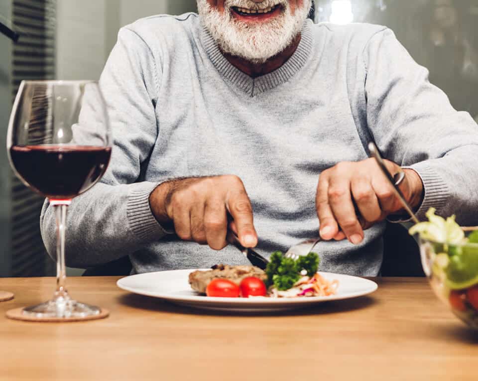 Senior man seating at a table cutting a piece of steak on a plate, next to a glass of red wine