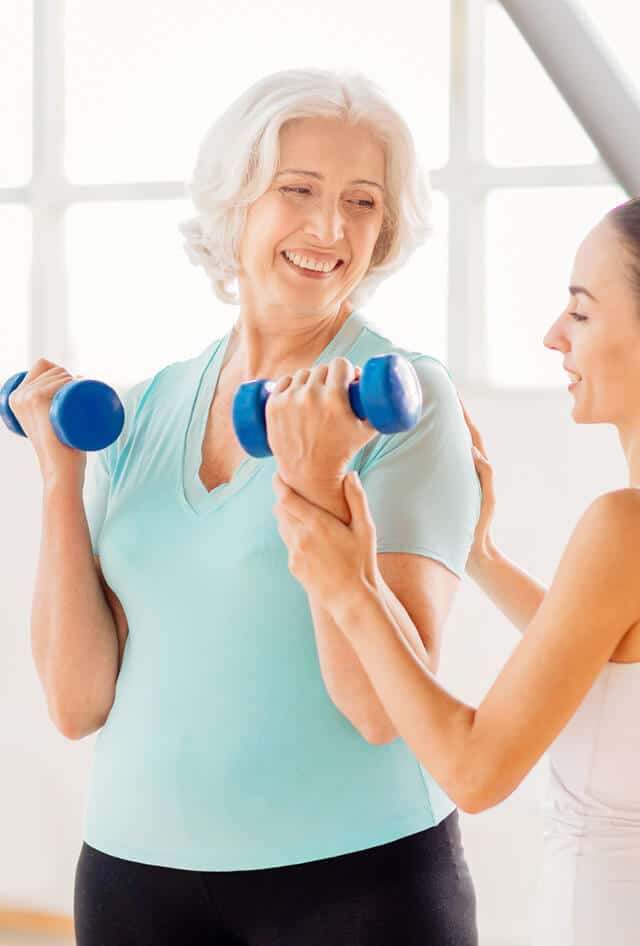 Older lady doing arm exercises with hand weights, assisted by a younger lady trainer